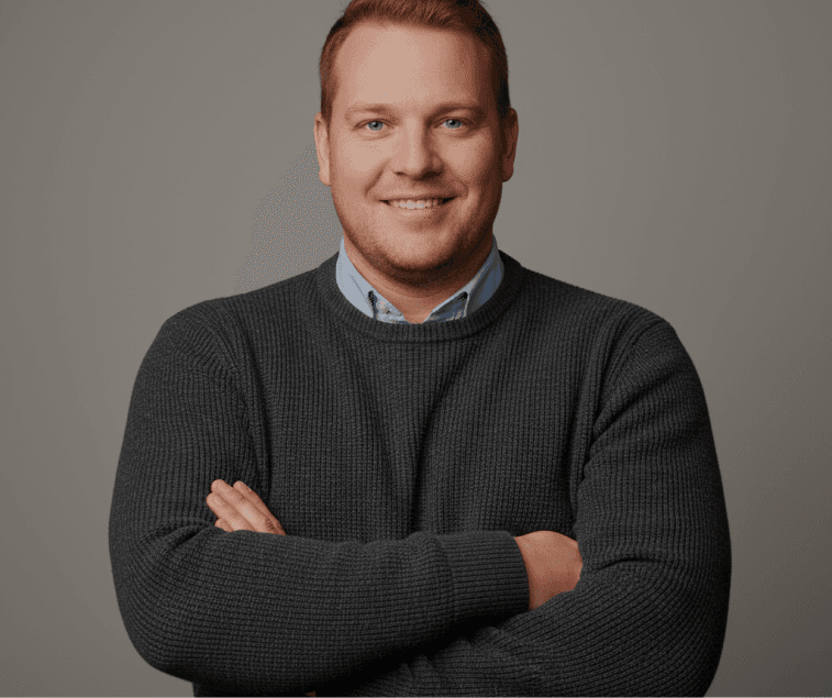Man in a grey knit sweater smiling, arms crossed